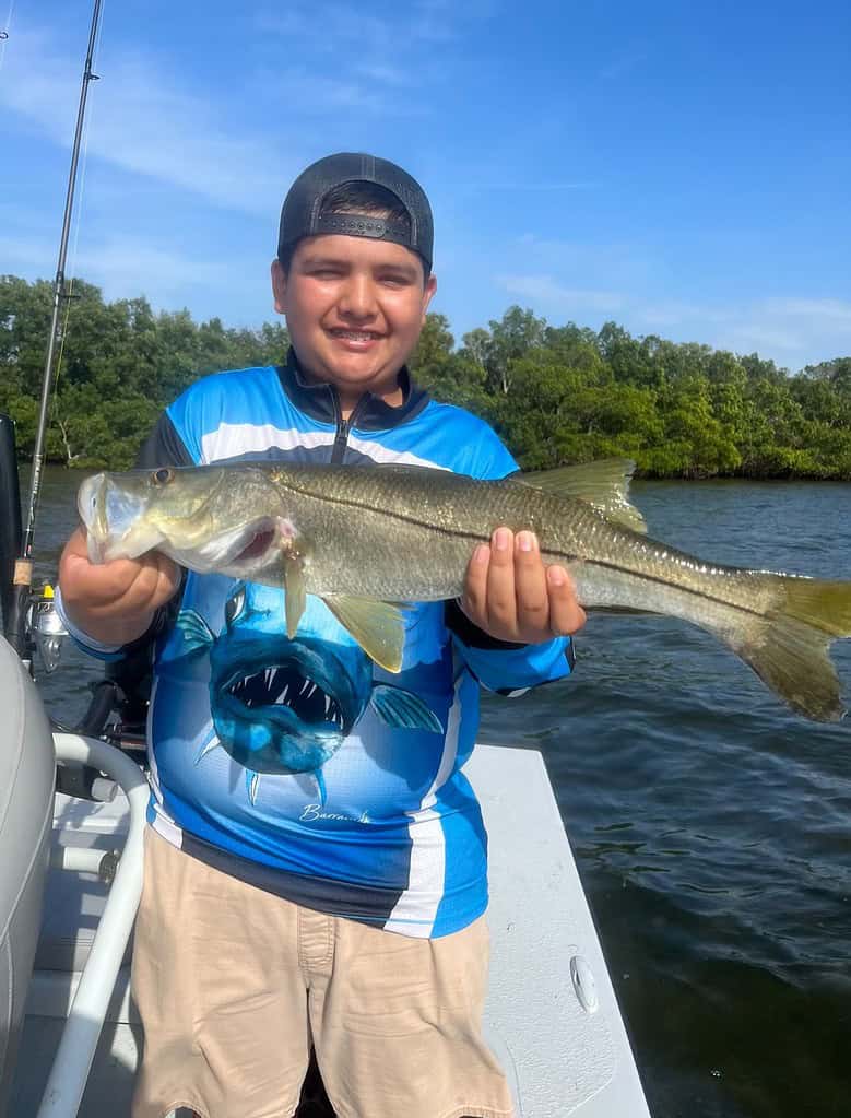 A young boy wearing a cap and blue fishing shirt, smiling and holding a large fish on a boat along the Florida gulf coast, with trees in the background.