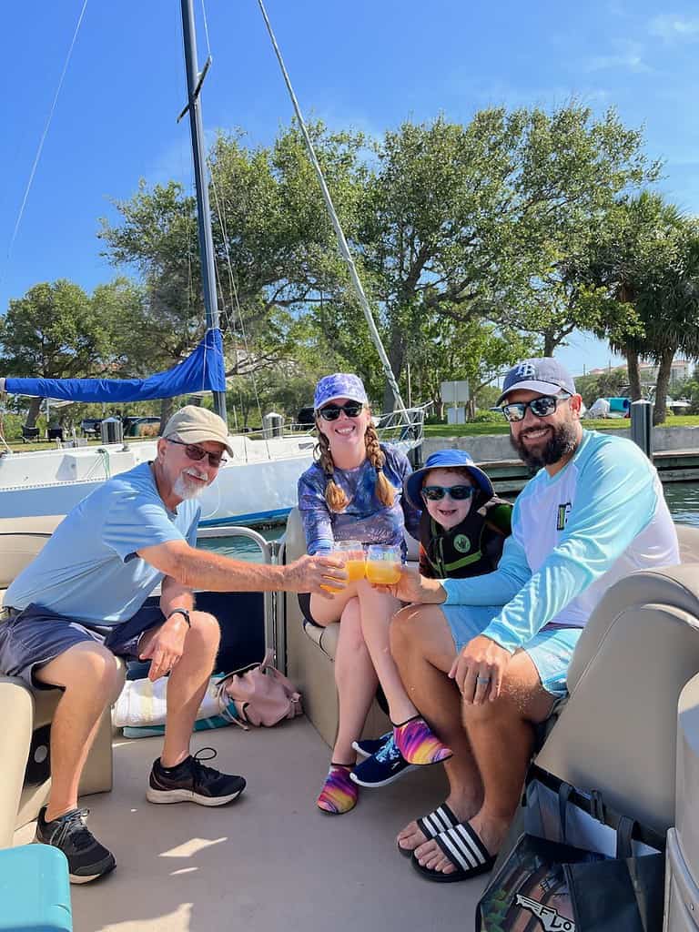 Two adults and two children smiling and sitting together on a boat in Siesta Key, holding a pineapple-shaped drink, with a river and sailboats in the background.