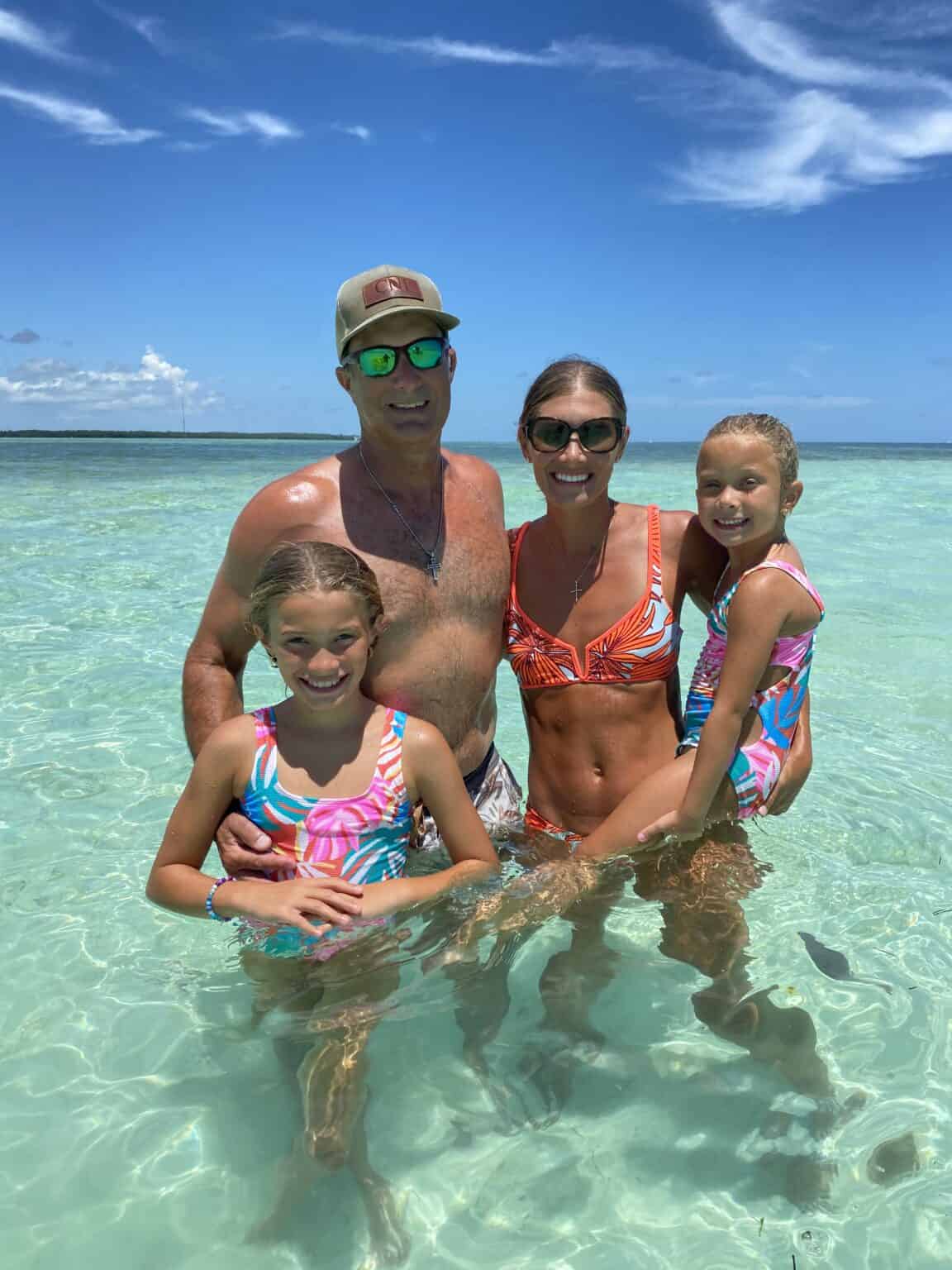 Family of four enjoying a sunny day in clear shallow water at a beach, smiling at the camera. two adults and two kids, all in swimwear.